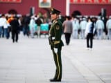 Ein chinesischer Militärpolizist steht auf einem Platz in Peking und überwacht das Geschehen. Im Hintergrund sieht man einige Menschen vor einem Gemälde von Mao Zedong stehen.