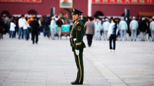 Ein chinesischer Militärpolizist steht auf einem Platz in Peking und überwacht das Geschehen. Im Hintergrund sieht man einige Menschen vor einem Gemälde von Mao Zedong stehen.