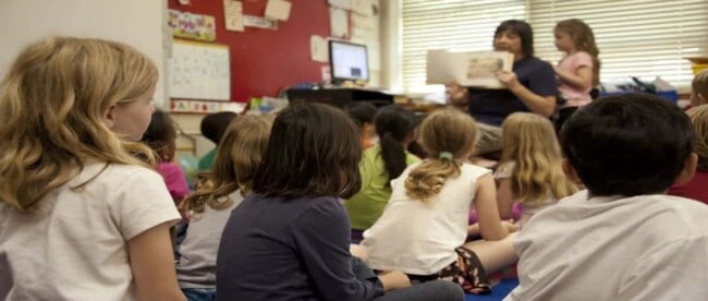 Schulkinder sitzen am Boden im Klassenzimmer und hören zu, während die Lehrerin ein illustriertes Buch vorzeigt.