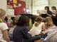 Schulkinder sitzen am Boden im Klassenzimmer und hören zu, während die Lehrerin ein illustriertes Buch vorzeigt.